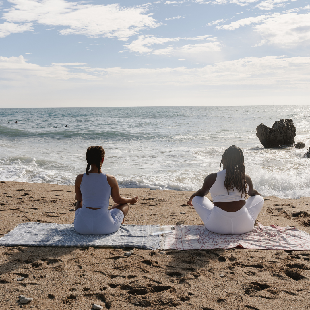 Women sitting on a beach on a blanket looking at the sea in a yoga sitting pose